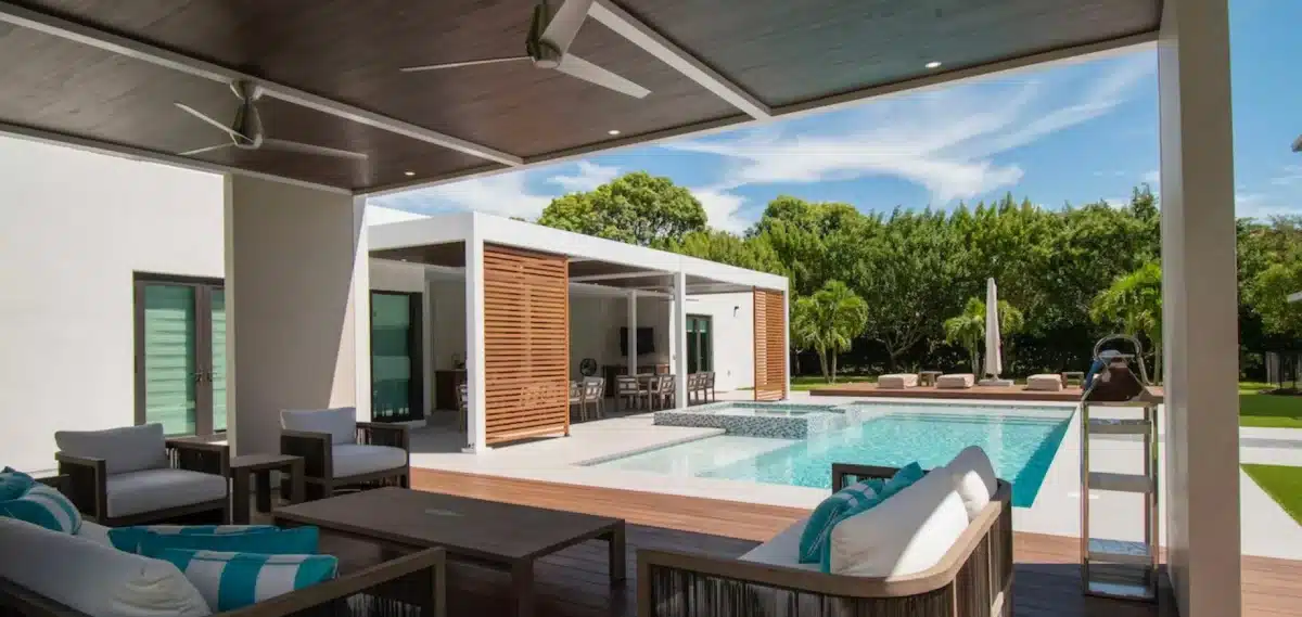 Outdoor living space featuring a covered patio with ceiling fans, modern seating, and a poolside lounge area with lush greenery and palm trees in the background.