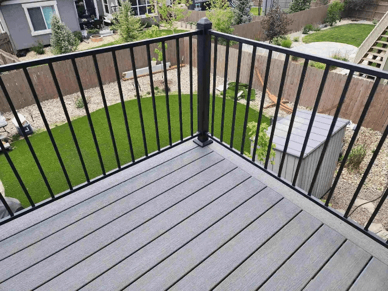 A gray composite deck with black metal railings overlooking a landscaped backyard with artificial turf and a wooden fence