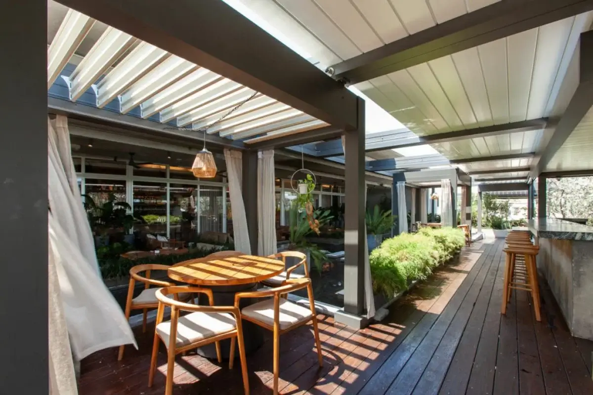 A stylish outdoor patio with a motorized louvered pergola, wooden decking, modern seating, hanging plants, and natural light filtering through adjustable roof panels