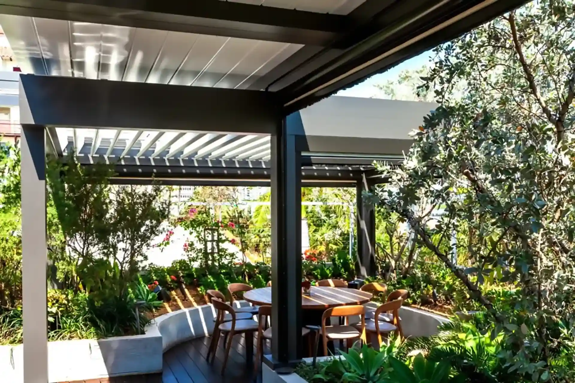 Modern outdoor dining area with a motorized louvered pergola, surrounded by lush greenery and vibrant landscaping.