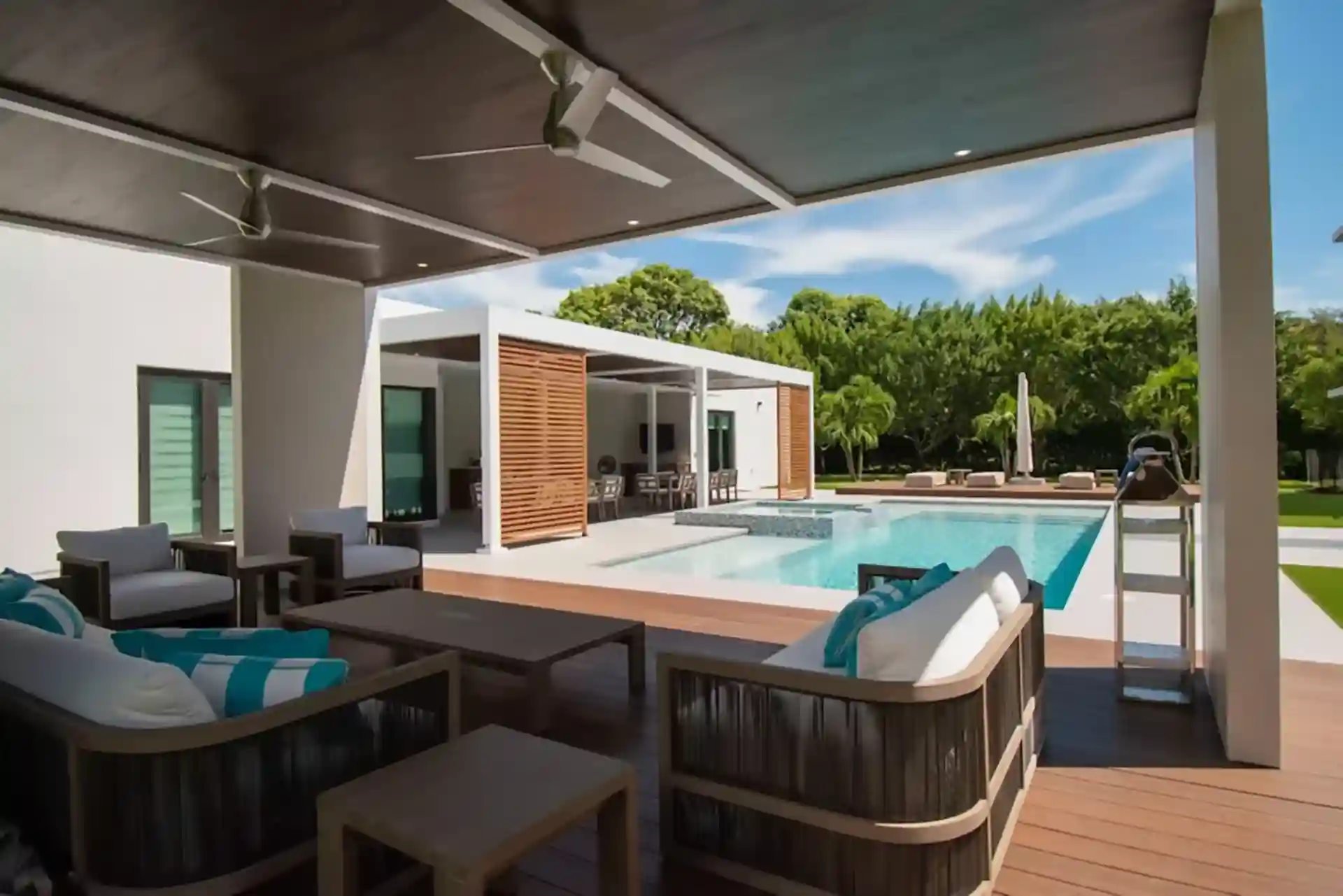 Luxury covered poolside deck with wooden flooring, modern outdoor seating, ceiling fans, pool view, and lush greenery