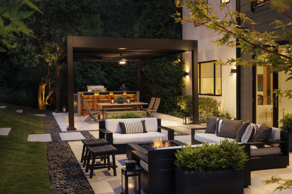Modern backyard patio with black aluminum pergola, outdoor kitchen bar, fire pit seating, and warm landscape lighting at night