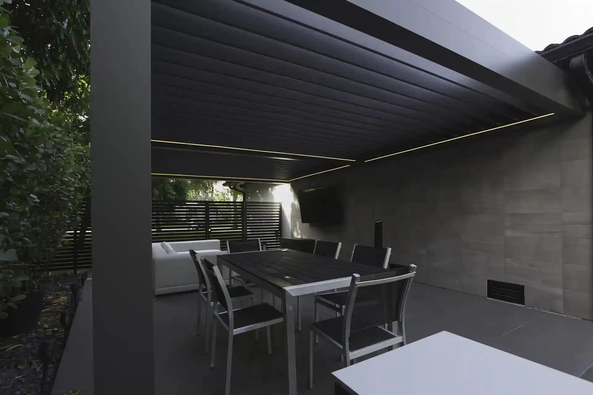 black-table-and-chairs-under-the-louvered-pergola-with-white-couch-in-front-of-the-tv-in-a-wall-featuring-black-privacy-screen-in-castle-rock-co