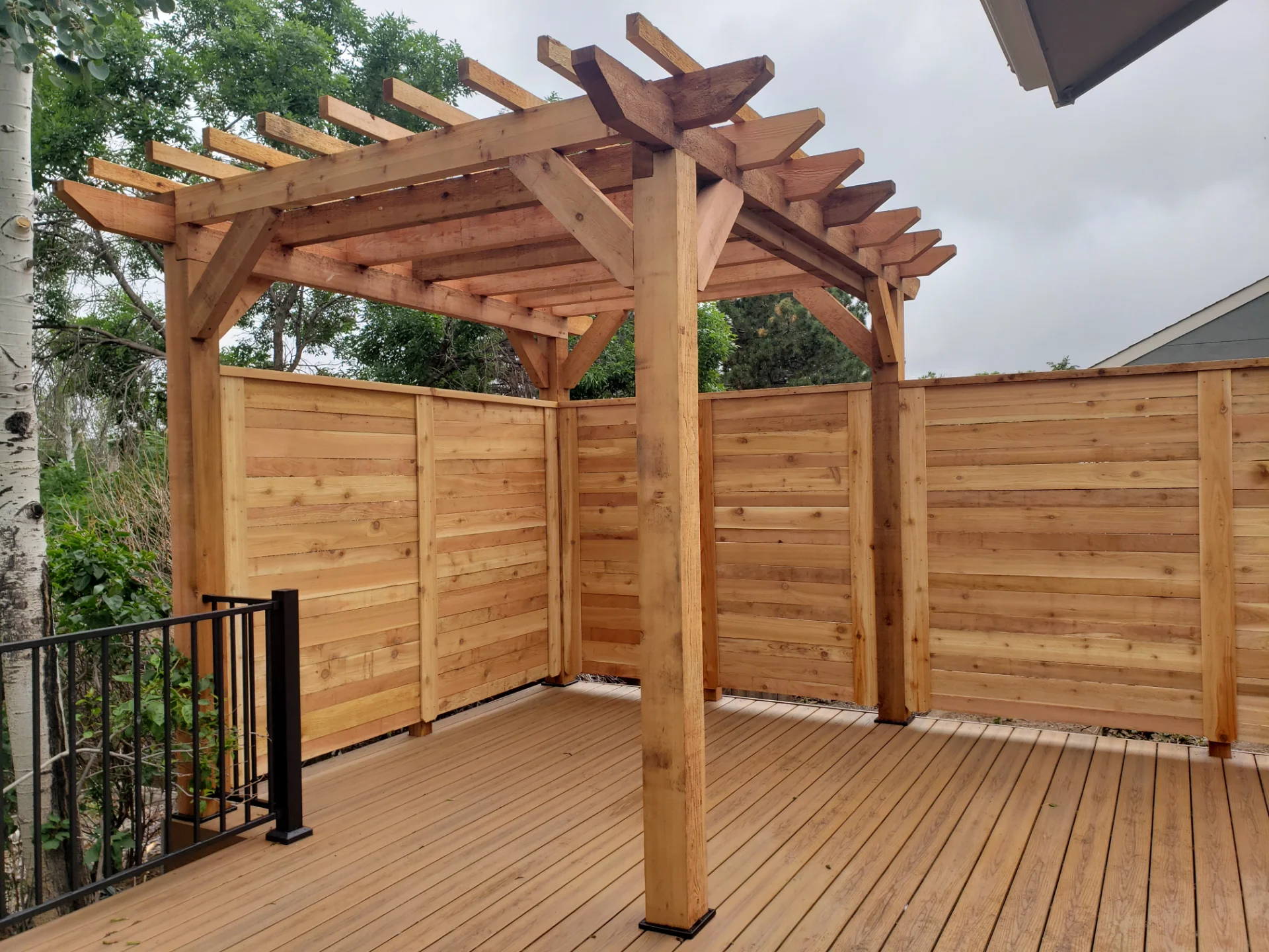 cedar-pergola-with-wooden-wall-screen-on-the-hard-wood-deck-with-black-metal-railing-featuring-trees-and-cloudy-sky-on-the-background-in-castle-rock-co