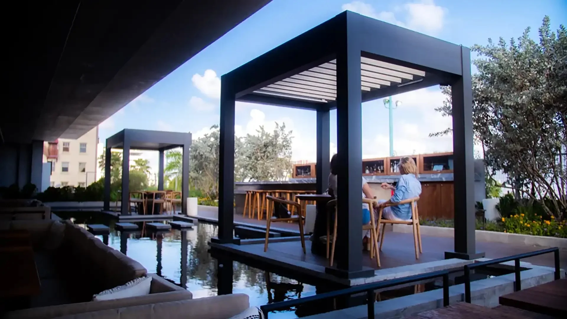 two-people-sitting-at-a-table-under-a-louvered-pergola-on-the-outdoor-bar-with-decorative-design-of-custom-deck-with-water-in-castle-rock-co